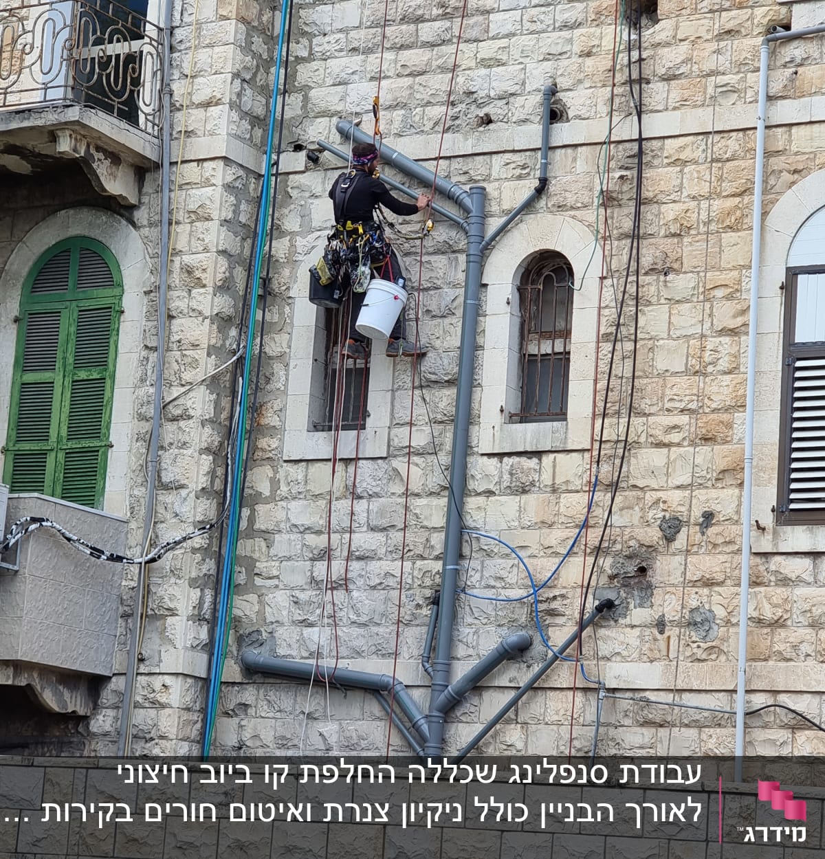 אדם תלוי על חבלים עם דלי וכלי עבודה ליד צינורות על קיר.